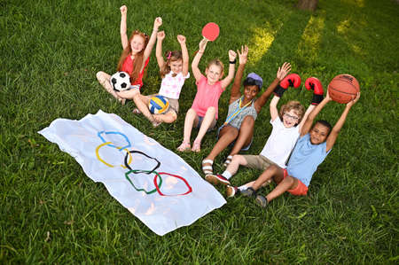 DONETSK, UKRAINE - JULY 10, 2021. Children of different nationalities with sports equipment in their hands are sitting on the grass against the background of a flag with Olympic symbols. Summer Olympic Games.の写真素材