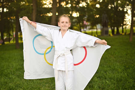 DONETSK, UKRAINE - JULY 10, 2021. A child girl in a white kimono holds a flag with Olympic symbols in her hands and smiles.の写真素材