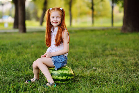 redheaded girl with watermelon grimaces and smiles sitting on the grass in the Parkの写真素材