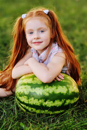 little funny girl with red hair leaning on a huge watermelon in the Park on the grass on a summer dayの写真素材