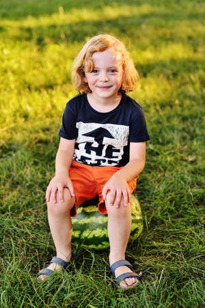 baby boy with curly hair sits on a big watermelon and smiles in the Park on a Sunny dayの写真素材