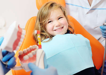 pediatric dentist shows how to properly brush her teeth to a little girl who is sitting in an orange...