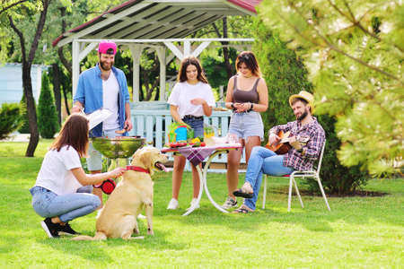 group of friends in the park at a picnic are barbecued, chatting, smiling and playing with a Labrador Retriever dog against the backdrop of greenery and a gazeboの写真素材