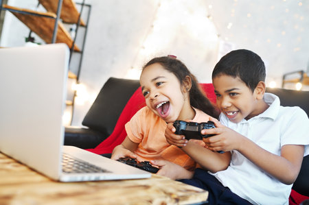 African American children - a boy and a girl holding a gamepad in their hands, play computer games on a laptop and laugh while sitting on the couch.の写真素材