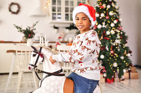 little African American boy in a gray Christmas sweater and a red santa hat smiles while sitting on a toy rocking horse against the background of a Christmas tree and a festive table.の写真素材