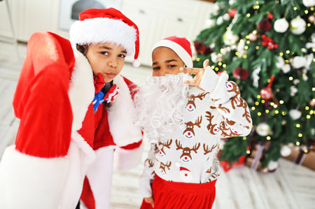 children - brother and sister in a large size santa costume with a false beard play and grimace against the backdrop of the Christmas tree.の写真素材