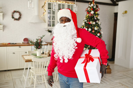 african american man in a red sweater with a white false beard and a santa hat smiles and holds a large white box with a red ribbon in his hands on the background of a christmas tree.の写真素材