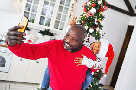 child in a sweater with deer and in a santa hat sitting on dads shoulders smiles and communicates with relatives via video link against the backdrop of a christmas tree.の写真素材