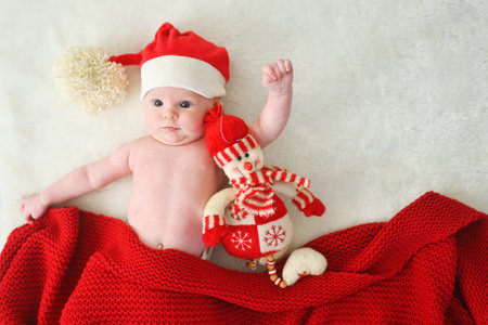 cute baby in a Santa hat with a big bubo covered with a red knitted blanket hugging a snowman toy on...