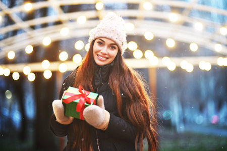 young woman in a knitted hat, who smiles and holds a Christmas present in her hands against the backdrop of snow and Christmas lightsの写真素材