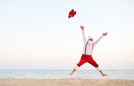 joyful Santa in red suspenders bounces barefoot against the background of the sea and the beach. Christmas vacation at the seaの写真素材