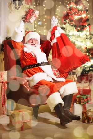 Santa Claus is smiling sitting on a red armchair and holding a bag of gifts against the background of a Christmas treeの写真素材