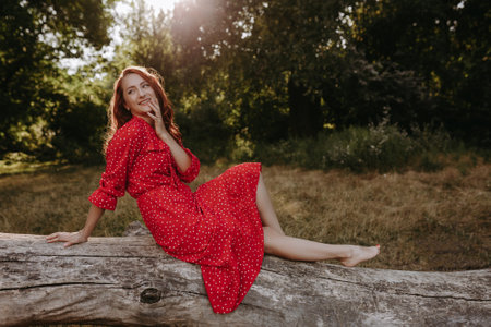 Pretty young adult woman in a red dress with white dots with red hair sitting on a dry fallen tree and posing on a camera. Female model touching her hair, head and faceの写真素材