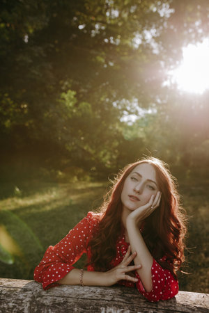 Portrait of pretty redhead woman in red summer loose shirt leaning by hands on a dry beam in wood...