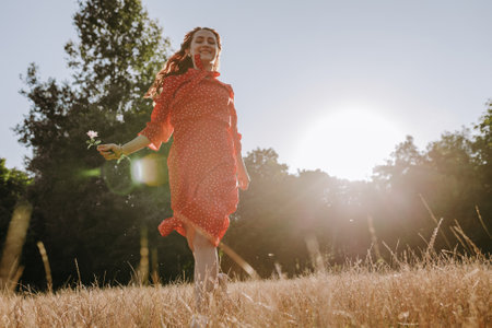 Pretty young redhead woman in red dress walking through meadow with flower in hand. Low angle view portrait of smiling female person on the forest bckgroundの写真素材