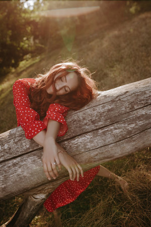 Young woman with red long curly thick hair lying down on a dry fallen tree in the middle of forest glade. Female person crossed her arms lying her head on treeの写真素材