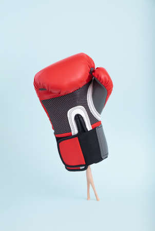 Plastic doll legs in heavy staggering boxing glove on minimal blue background. Art concept.