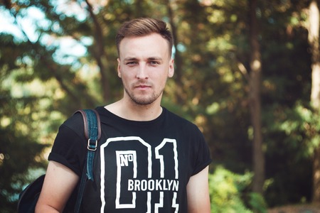 Portrait of a stylish young man in a black shirt with a backpack on his shoulders. A student in the park.の写真素材