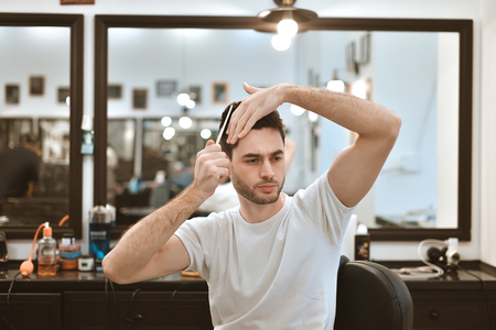 Working process in modern barbershop. Handsome hairdresser serving customer, styling hair for male client using hairdryer. Side view portrait of attractive young man getting trendy haircut. Indoorsの写真素材