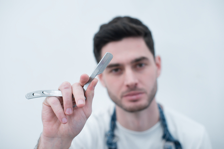 Barber keeps the razor against the background of a white wall. Bearded brutal young man with tools in his hands. Focus on tools.の写真素材