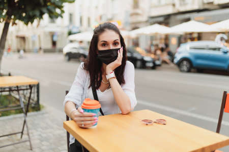 Young woman sitting on the street in a mask with a cup of coffee.の写真素材