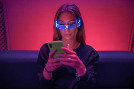A young woman is sitting on the couch wearing neon glasses and using a gadget.の写真素材