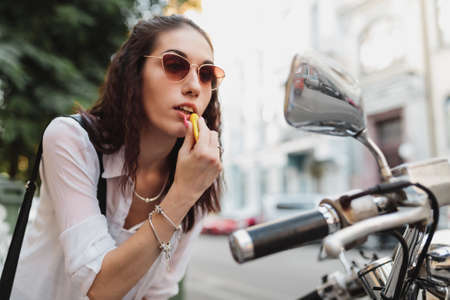 Young woman paints her lips looking in the mirror of a motorcycle.の写真素材