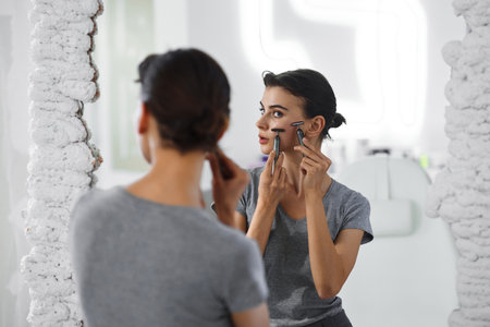 A young woman massages her face with gouache while looking in the mirror.の写真素材
