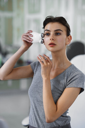 A young woman massages her face with a microcurrent gadget while looking in the mirror.
