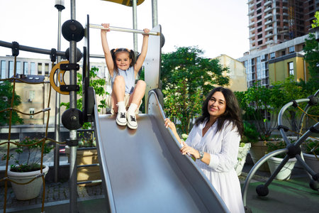 A mother watches her daughter play on a slide in a sunny urban playground during the afternoonの写真素材
