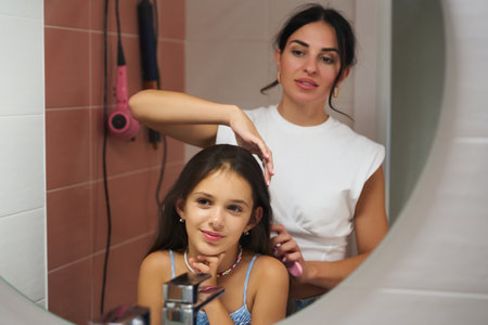 Mother brushing her daughters hair in a bathroom with soft lighting and pastel-colored tilesの写真素材