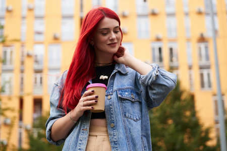 Woman with vibrant red hair stands against a yellow wall holding coffee