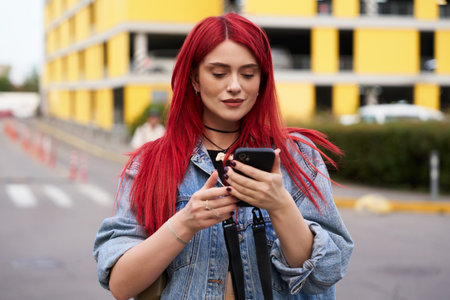 Young woman with red hair using smartphone on urban street in daylightの写真素材