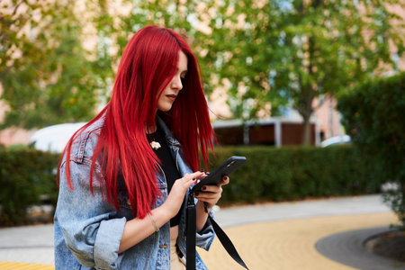 Young woman with vibrant red hair using smartphone in urban park setting