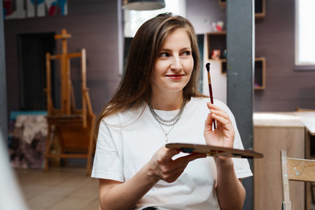 Young woman with a paint palette smiles while preparing to create art indoorsの写真素材