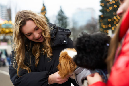 Smiling girl holds fluffy dog while chatting with a friend outdoors during winter festivities