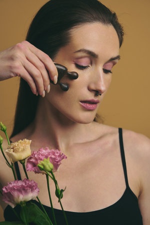 Woman using facial roller on her cheek with flowers in the backgroundの写真素材