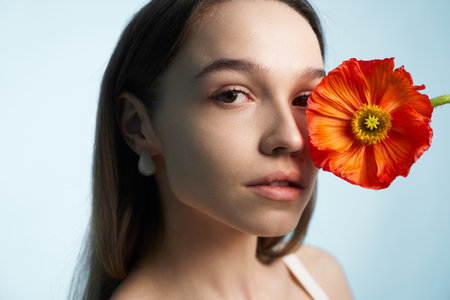 Young woman poses closely with a vibrant orange flower against pastel backgroundの写真素材