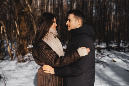 Couple embracing warmly in a snow-covered forest during winter sunlightの写真素材