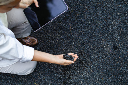 Close up of woman examines a pile of dark crushed stone while holding a tablet during an outdoor site visit. Concept of material evaluation, engineering inspection and infrastructu