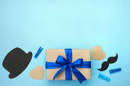 Fathers day composition. Gift box wrapped in karft paper with blue ribbon, hearts, mustache, black hat and pins on blue background. Top view flat lay with free space for text. Greeting card.の写真素材
