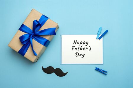 Happy Fathers day composition. Gift box with blue ribbon, mustache and signed card on blue background. Top view flat lay. Greeting card for dad.の写真素材