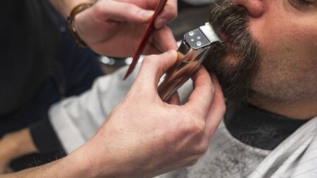 Bearded man in barbershop. Haircut concept. Man visiting hairstylist in barbershop. Barber works with hair clipper. Hands of barber with hair clipper, close up.の写真素材