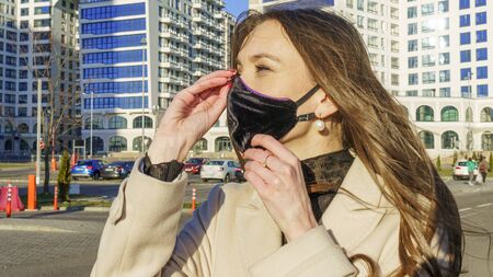Coronavirus fashionable medical face mask worn by young female with dark long hair, standing at tram stop on a urban city street on dusk. Stop covid 19 pandemic or air pollution concept.の写真素材