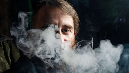 Brutal man with large beard and fashionable haircut smokes an electronic cigarette or hookah on dark background.の写真素材