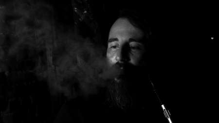 Brutal man with large beard and fashionable haircut smokes an electronic cigarette or hookah on dark...