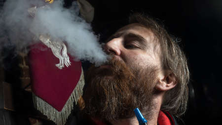 Close-up photo of man smoking traditional hookah pipe. Man exhaling smoke in hookah cafe on dark...