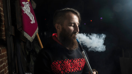 Brutal man with large beard and fashionable haircut smokes an electronic cigarette or hookah on dark...