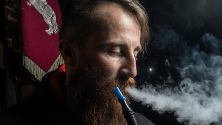 Brutal man with large beard and fashionable haircut smokes an electronic cigarette or hookah on dark...