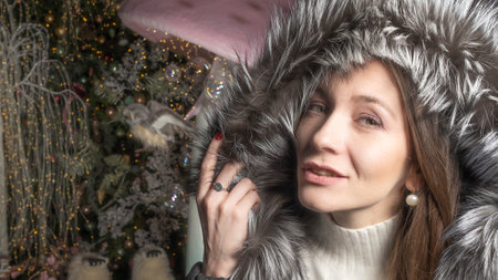 The caucasian white woman with black hair and jacket with fur hood stands against background of the Christmas tree with the bright lights.の写真素材
